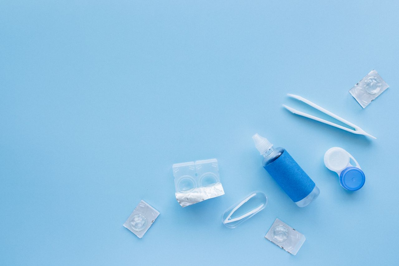 Services Flat lay of contact lens care kit with solution and tweezers on blue backdrop.